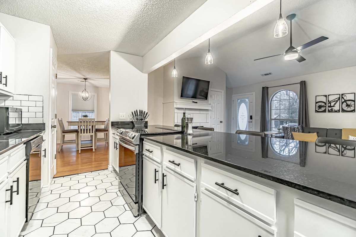 The kitchen features white cabinetry with sleek black countertops and a geometric tiled floor. A window allows natural light to illuminate the space. The open layout connects to the dining area visible in the background, showcasing a light fixture and wooden dining table.