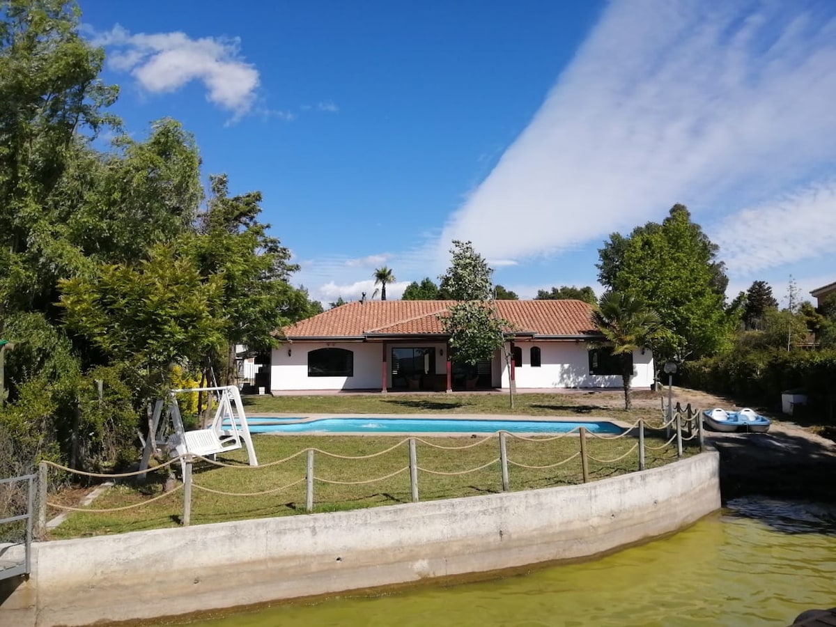 A spacious house is set against a clear blue sky, featuring a swimming pool bordered by a neatly maintained lawn. Lush trees and landscaping are visible around the property, with a calm lake in the foreground, reflecting the serene environment.