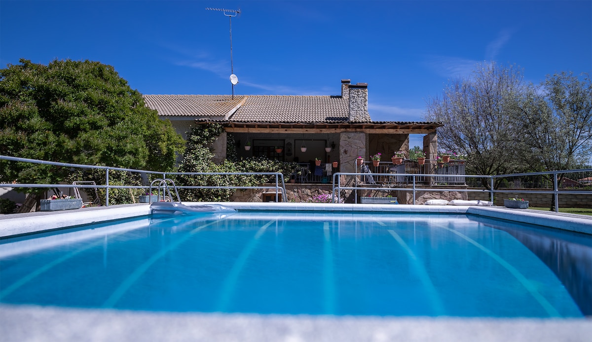 A clear swimming pool reflects the blue sky, with the house's stone façade visible in the background. Lush green foliage surrounds the area, providing a sense of privacy and tranquility.