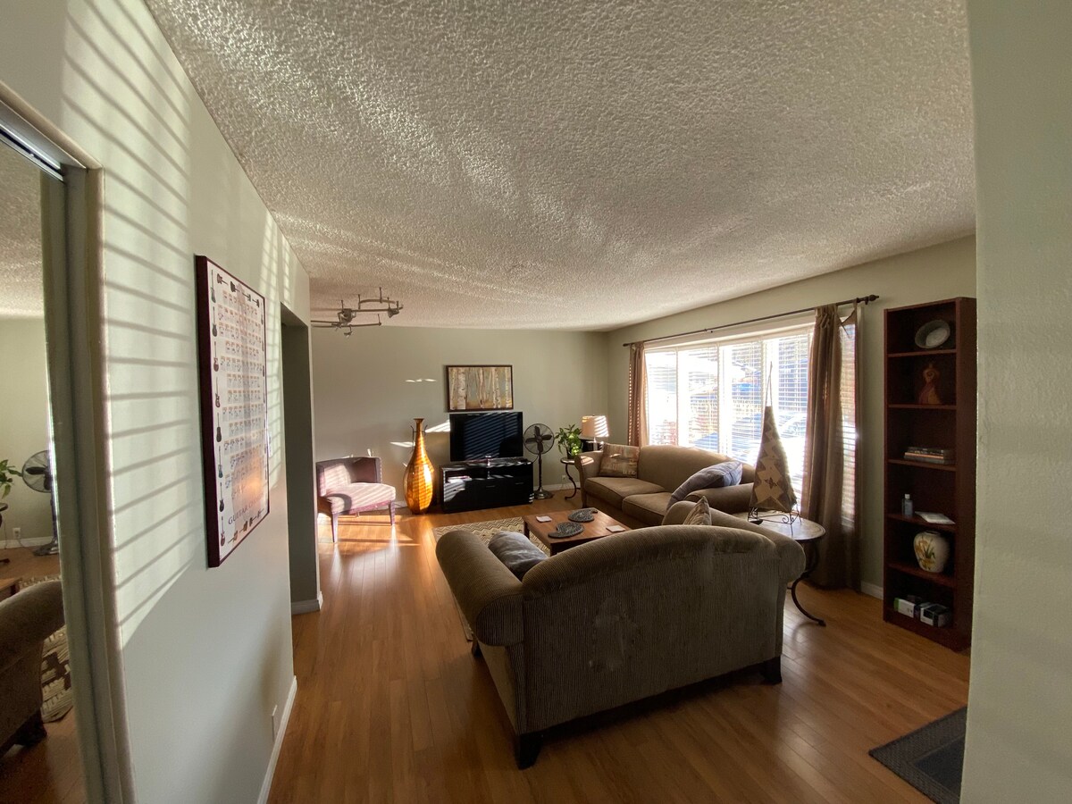 A spacious living room is filled with natural light, featuring comfortable seating arranged around a coffee table. A television sits on a black console, while large windows adorned with soft curtains offer a view of the outside. A decorative bookshelf adds character to the room.