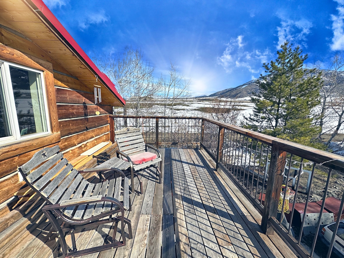 A wooden deck extends from the cabin, furnished with two rustic chairs and a lounge chair. The expansive view showcases surrounding mountains and a clear sky. Sunlight casts dynamic shadows across the deck, highlighting the planked flooring.