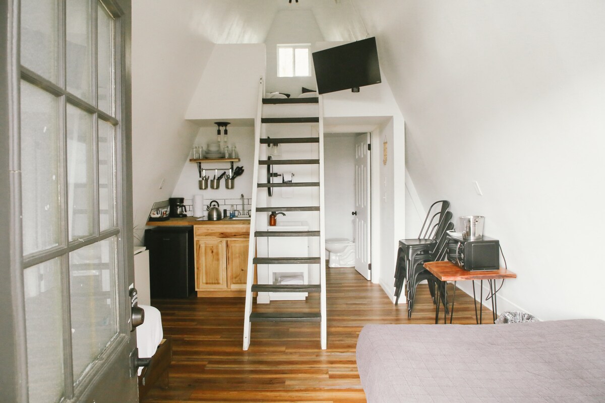 The interior of the A-Frame cabin is captured, highlighting the open layout featuring a staircase leading to the loft. A kitchenette with wooden cabinetry is visible on the left, complemented by a dining table with chairs and a wall-mounted TV above the stairs.