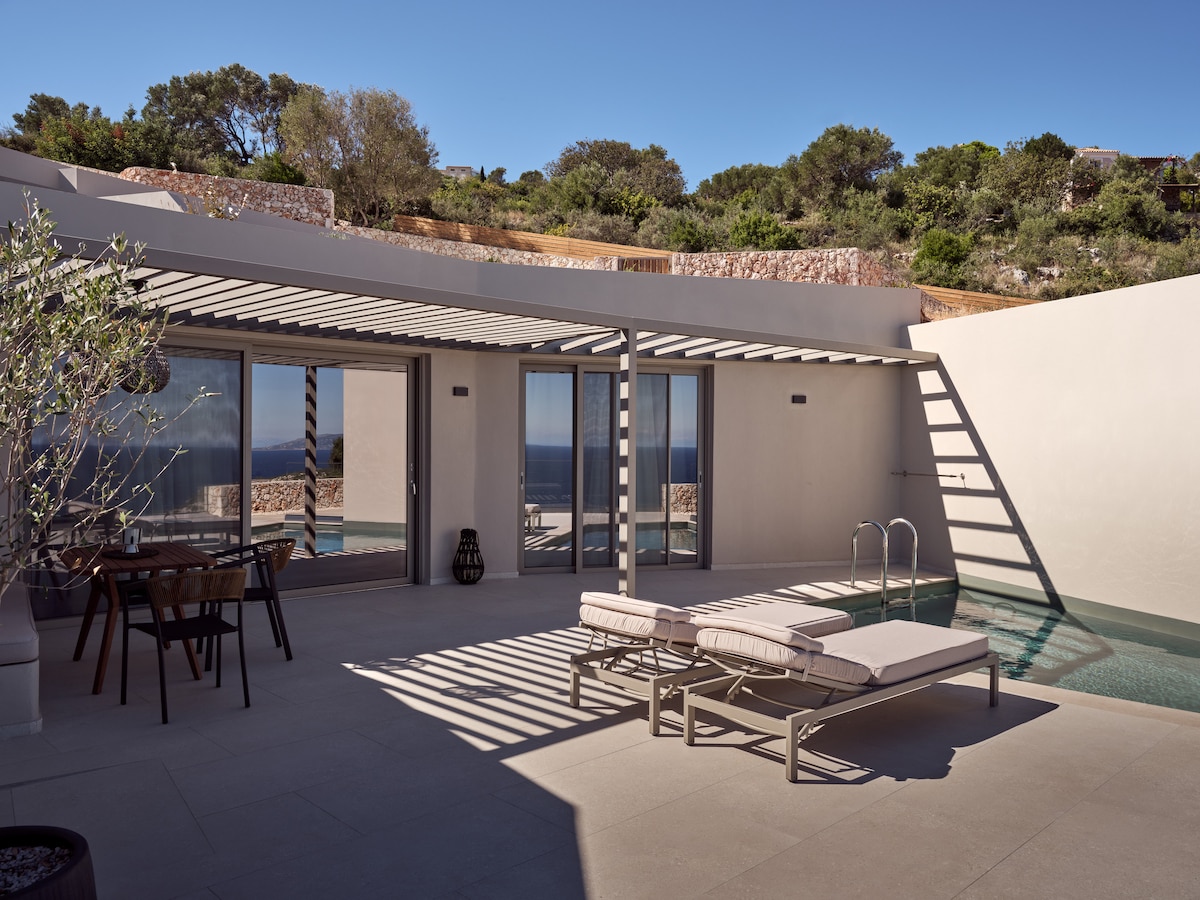 A private patio features two sunbeds beside a shimmering pool, with views of the Ionian Sea and surrounding Mediterranean landscape. Glass doors provide access to the interior, and a dining table is positioned for outdoor meals under a shaded pergola.