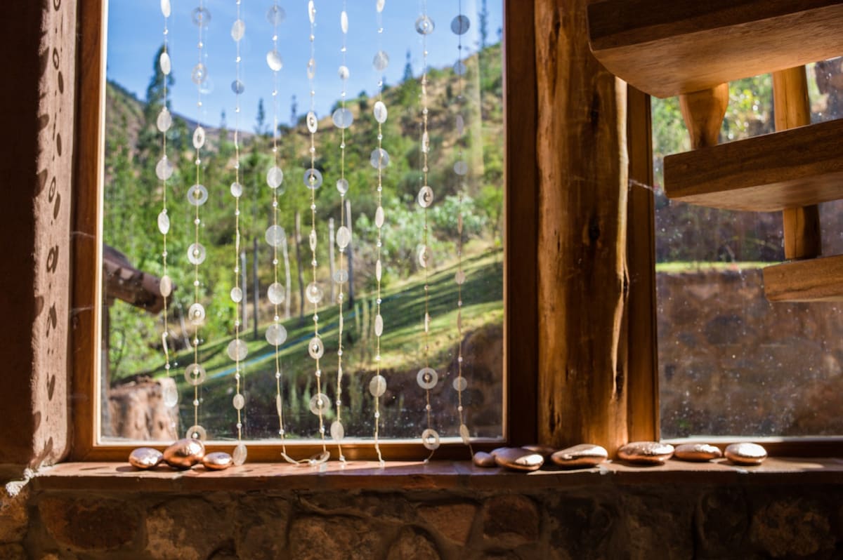 A wooden window frames a view of lush greenery outside, with small decorative stones arranged along the windowsill. Delicate beads hang in the window, allowing light to filter through and create a soft glow in the interior space.