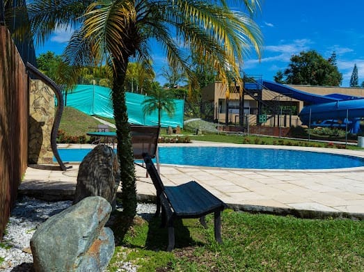 A large, inviting swimming pool is surrounded by lush greenery and palm trees. A wooden lounge chair rests on the grass, while a waterslide and play area are visible in the background, enhancing the recreational atmosphere of the space.