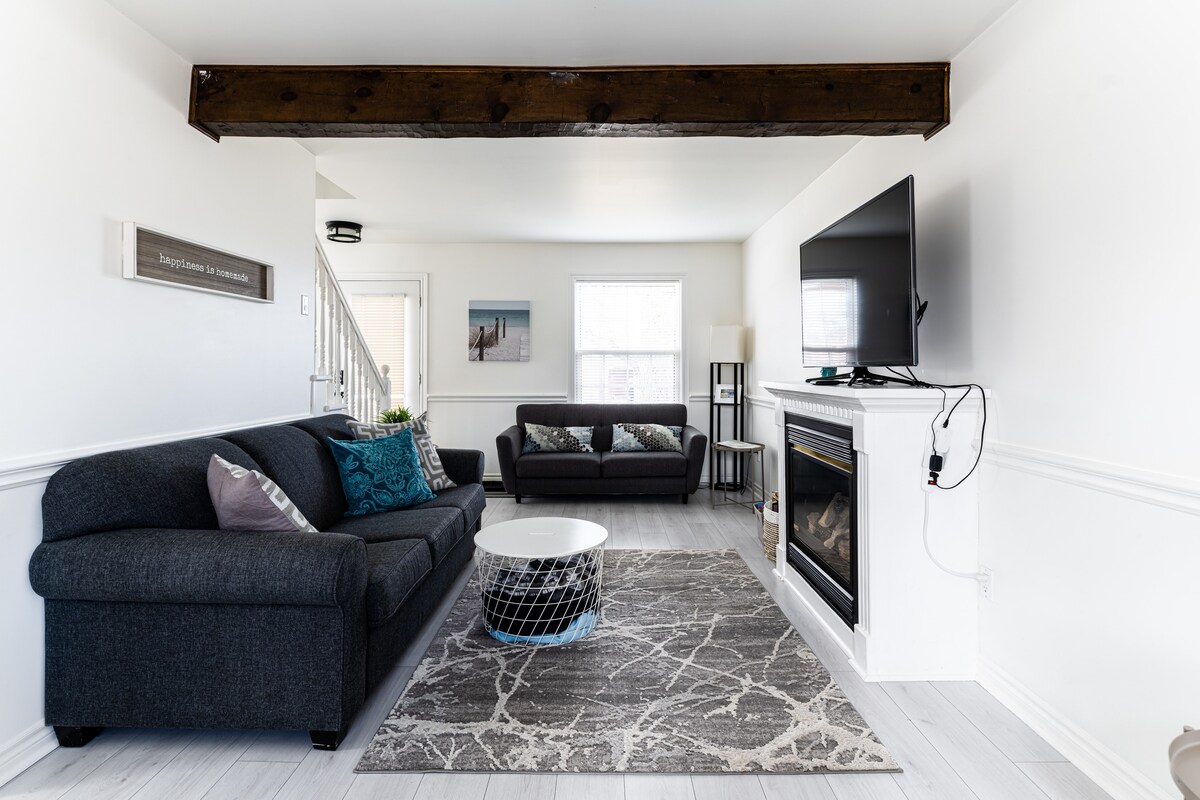A cozy living room is present, featuring two sofas, a coffee table, and a large Smart TV mounted above a fireplace. Natural light enters through the window, illuminating the light-colored walls and the soft gray area rug on the floor.