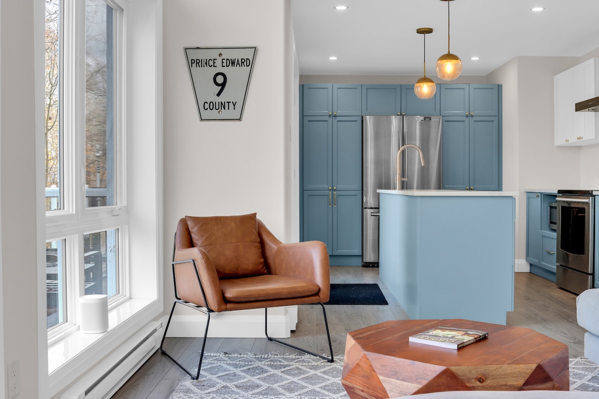 A modern kitchen and living area are displayed, featuring a light blue kitchen island and cabinetry. A brown leather armchair is positioned by a large window, with a coffee table centered in the space. Warm pendant lights enhance the inviting layout.