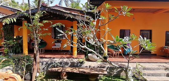 The exterior of the villa is presented with vibrant orange walls and lush greenery. A wooden bench is positioned along a stone pathway surrounded by tropical plants. Seating areas are visible on the porch, providing inviting spaces to relax in the shade.