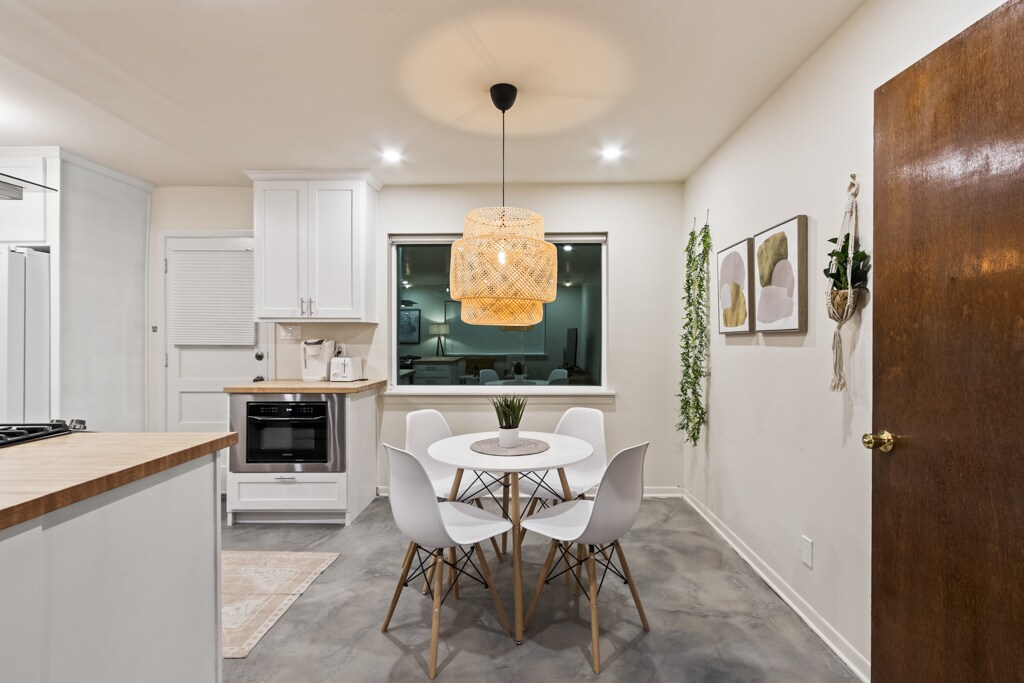 A dining area features a round white table surrounded by four modern chairs. A statement light fixture hangs above, casting a warm glow. A large window presents an outdoor view, while subtle greenery is displayed on the walls, enhancing the minimalist decor.