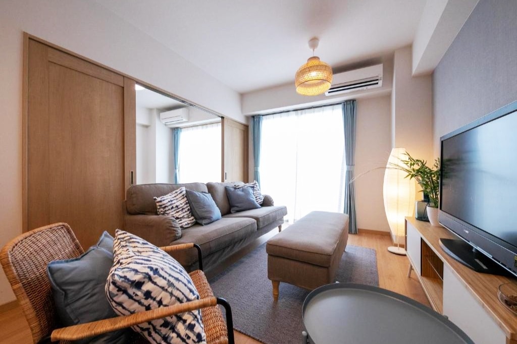 A spacious living area features a comfortable sofa adorned with decorative pillows. A light wood coffee table is positioned on a soft area rug, with natural light streaming through large windows adorned with light blue curtains. A modern lamp adds a cozy touch.