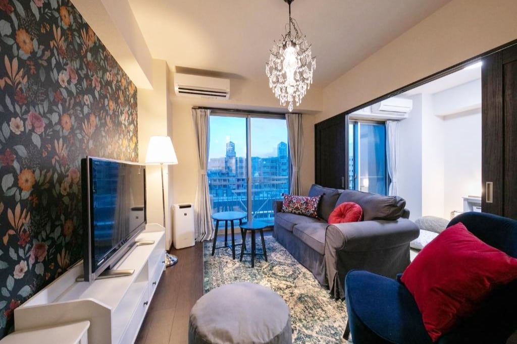 A cozy living area features floral wallpaper and a spacious couch with colorful throw pillows. A television sits on a white unit, while a small table and ottoman are positioned nearby. Large windows allow natural light to fill the space, revealing city views.