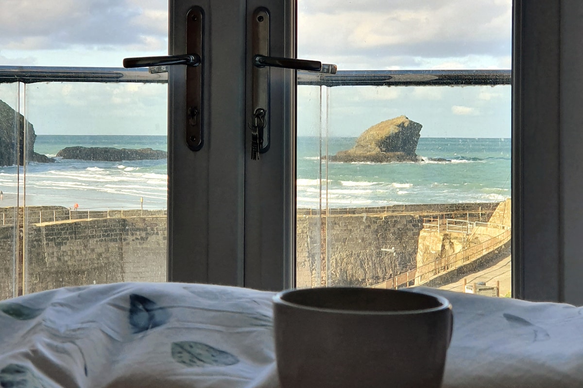 The image showcases a serene view through glass doors, capturing the ocean waves against a rocky shoreline. A cozy set of sheets is slightly visible in the foreground, accompanied by a simple cup, suggesting a relaxed morning setting with nature at the doorstep.