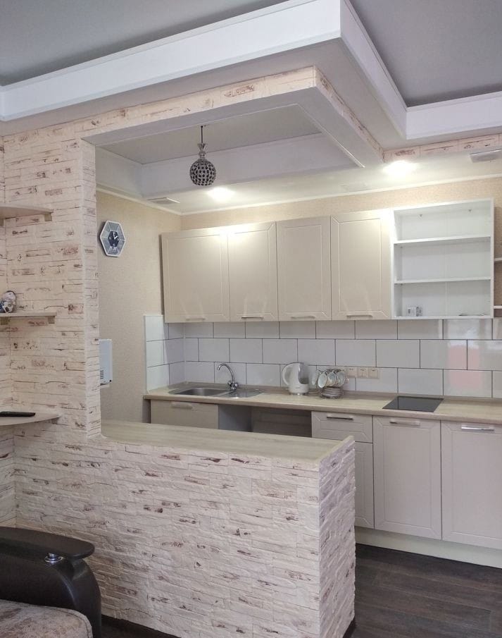 A compact kitchen area features modern cabinetry and a partial stone wall. A sink and countertop provide workspace, while dishware and appliances are visible. Soft overhead lighting enhances the functional design, contributing to the overall practicality of the space.