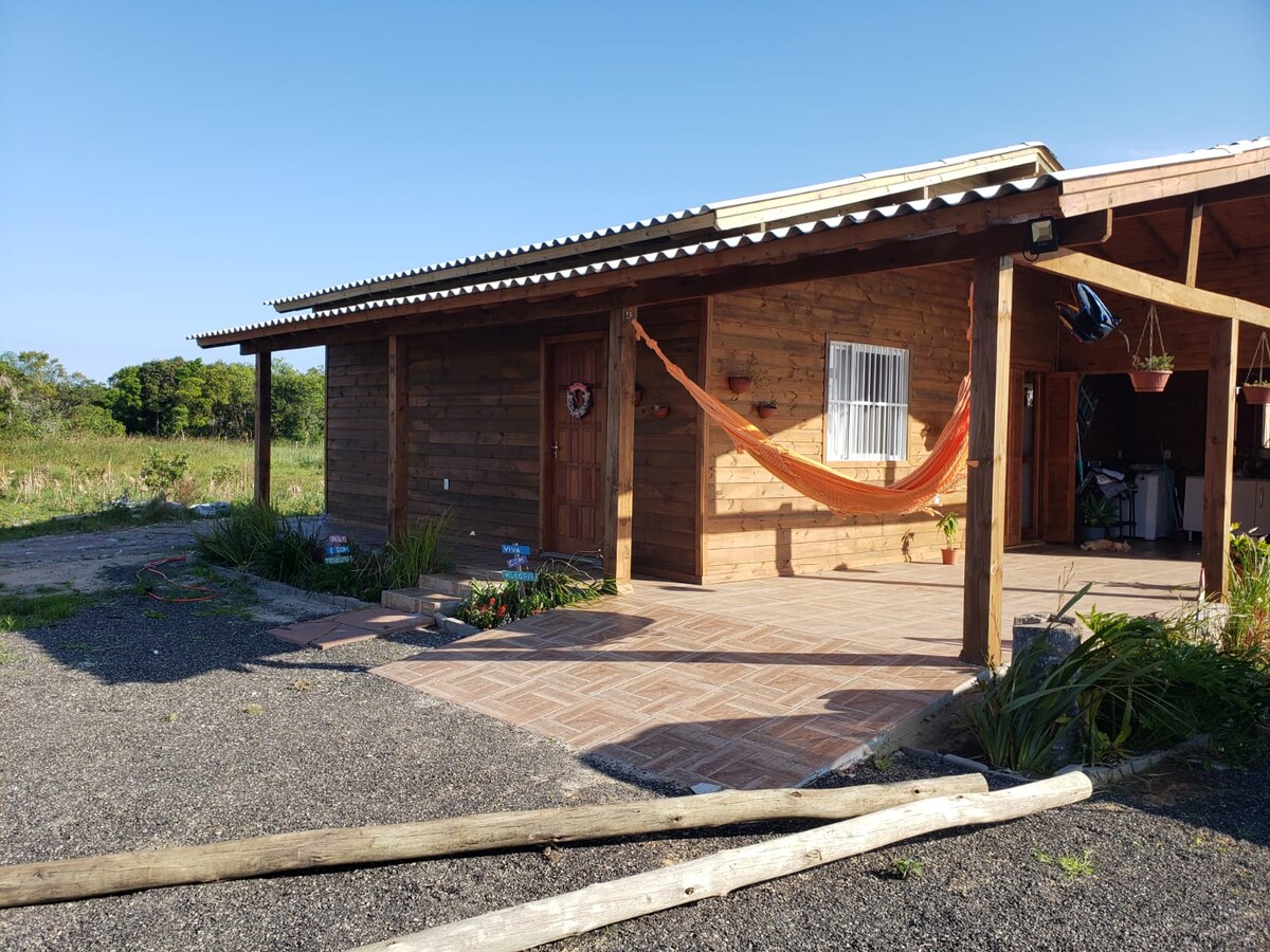 A wooden exterior of a single-story house is presented, with a welcoming porch featuring a colorful hammock. Surrounding greenery and gravel pathways lead to the entrance, while a bright blue sky adds to the serene atmosphere of the outdoor space.