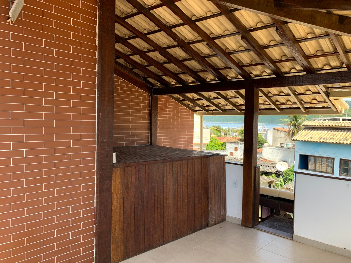 An open terrace space is presented, featuring a wooden bar area. The ceiling is supported by exposed beams, providing a spacious feel. In the background, a glimpse of the ocean can be seen, enhancing the inviting environment.