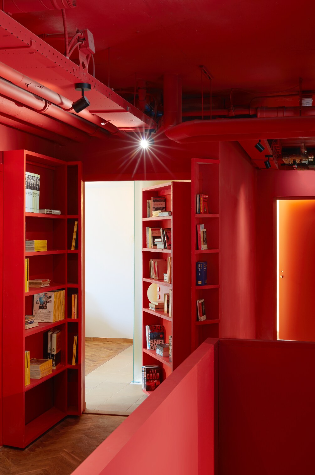 A vibrant corridor features walls painted in red, with built-in bookshelves lining the sides. Brightly colored books are neatly arranged on the shelves, leading to a doorway at the end. Soft lighting illuminates the space, creating a unique and inviting atmosphere.