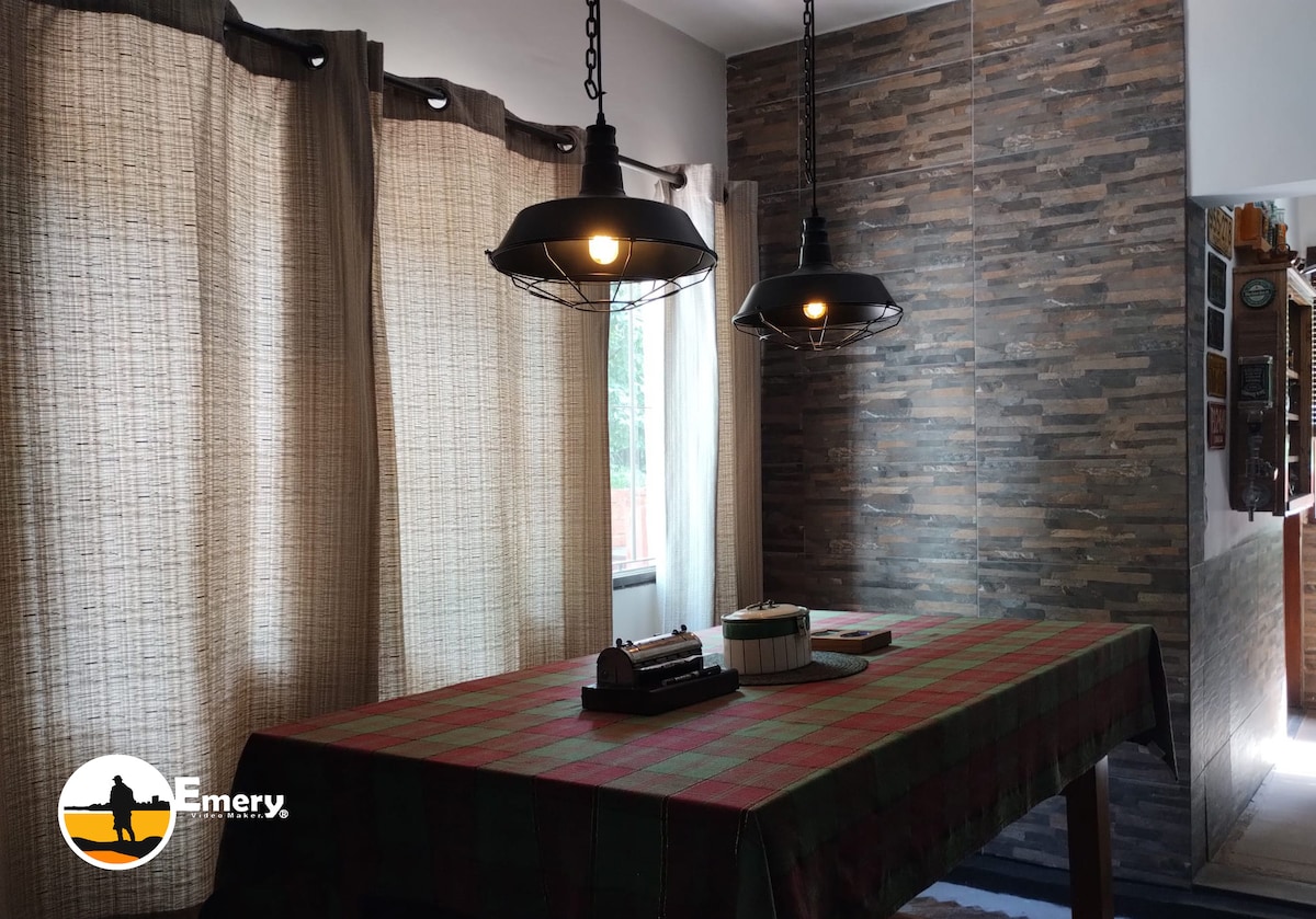 A dining area features a spacious table covered with a patterned cloth, surrounded by natural light from large windows draped with light curtains. Industrial-style pendant lights hang above, adding a modern touch to the warm atmosphere.