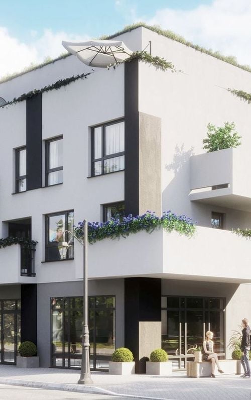 A modern building features a minimalistic design with clean lines and large windows. Decorative plants are positioned along the exterior, enhancing the welcoming appearance. A small seating area is visible at the ground level, providing a space for relaxation.