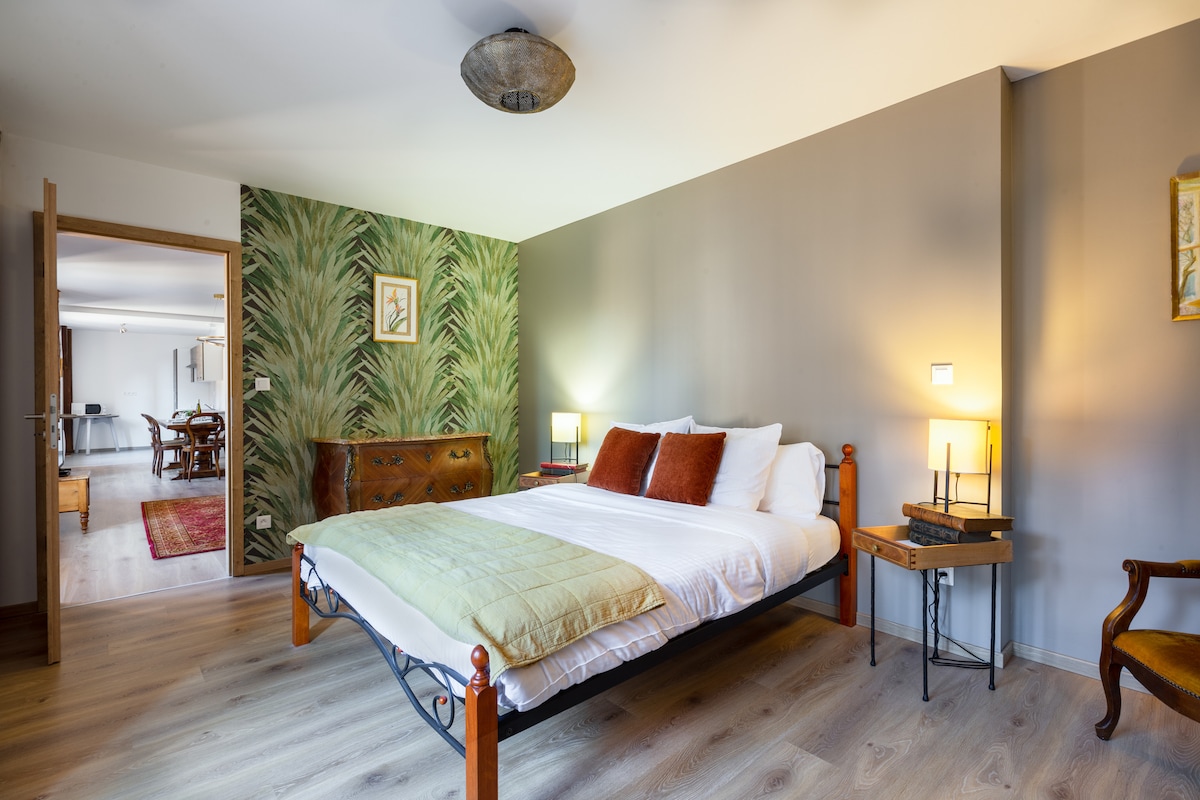 This spacious bedroom features a comfortable bed with decorative pillows, complemented by a soft green throw. A wall adorned with a leafy pattern adds character, while warm lighting from bedside lamps creates a cozy ambiance. A wooden chair and dresser enhance the room's charm.