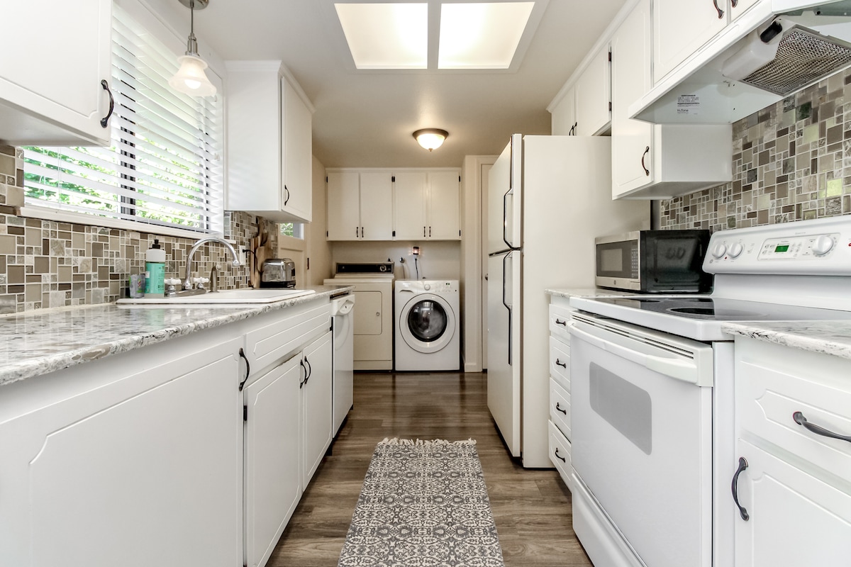 The fully equipped kitchen features modern appliances, including a refrigerator, stove, and microwave. A washer and dryer are conveniently installed, and a decorative rug runs along the floor. Natural light enters through partially open blinds, illuminating the neutral color scheme and tiled backsplash.