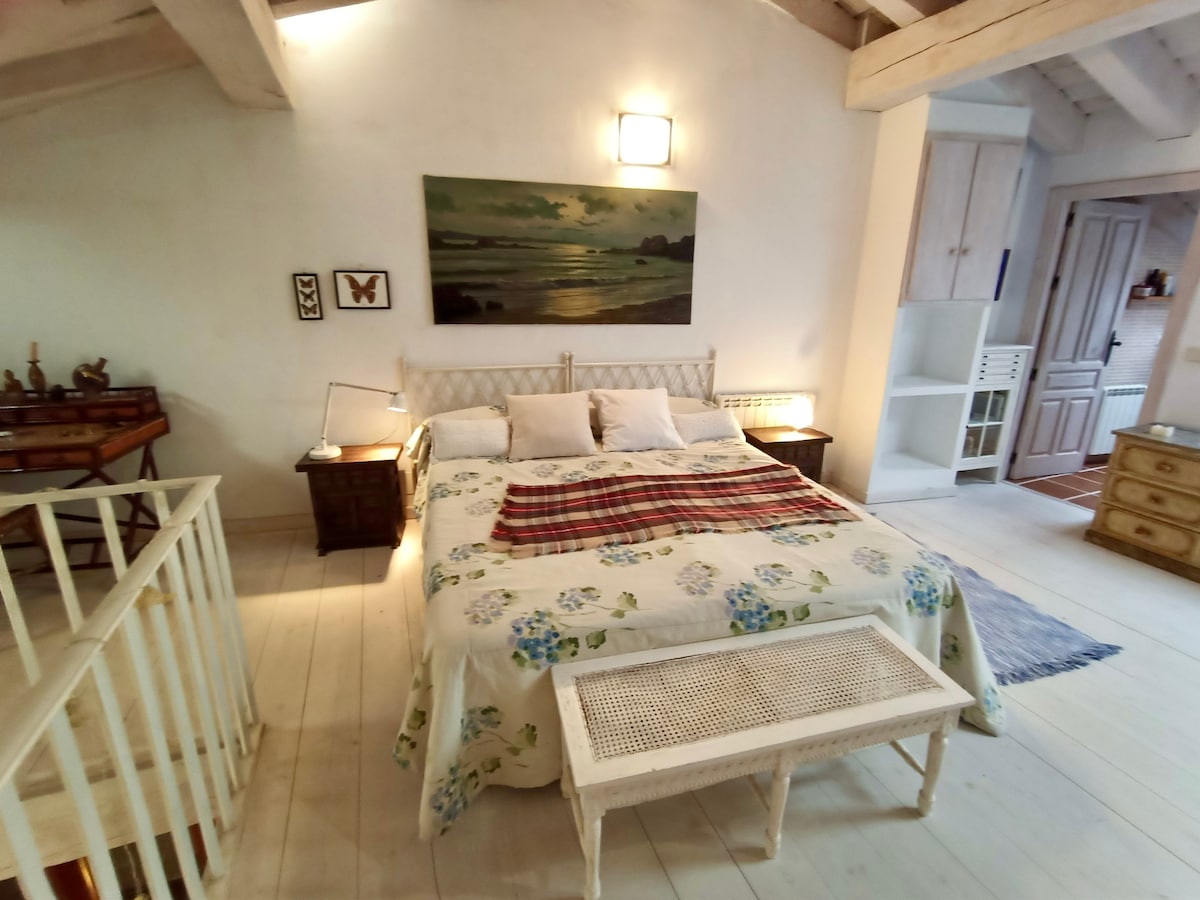 A spacious bedroom features a comfortable bed dressed in floral-patterned linens. Soft lighting illuminates the area, highlighting decorative accents like a wall-mounted painting and a small table at the foot of the bed. The room has a rustic charm with wooden beams and light-colored floors.