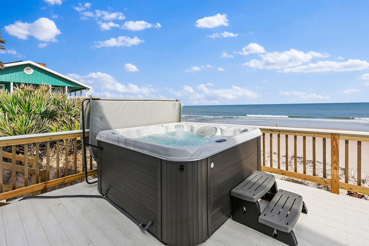 A hot tub is positioned on a sunlit deck, offering views of the ocean and sandy beach beyond. Surrounding foliage adds to the relaxing ambiance. Clear skies and gentle waves create a peaceful coastal atmosphere.