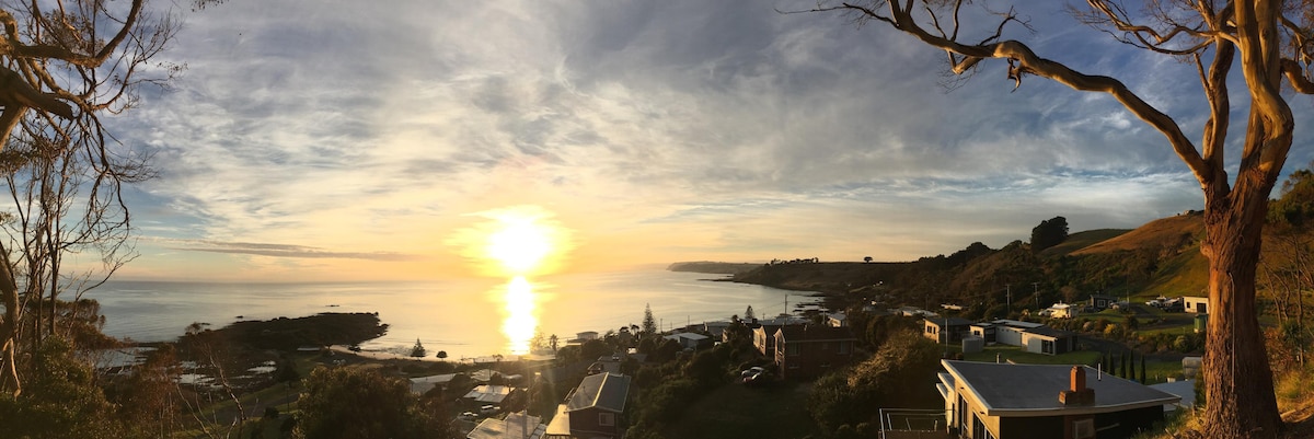 A panoramic view captures a serene sunrise over the ocean, with soft hues of gold and blue across the sky. Coastal homes and lush greenery are visible along the shoreline, reflecting the tranquil atmosphere of Boat Harbour Beach.