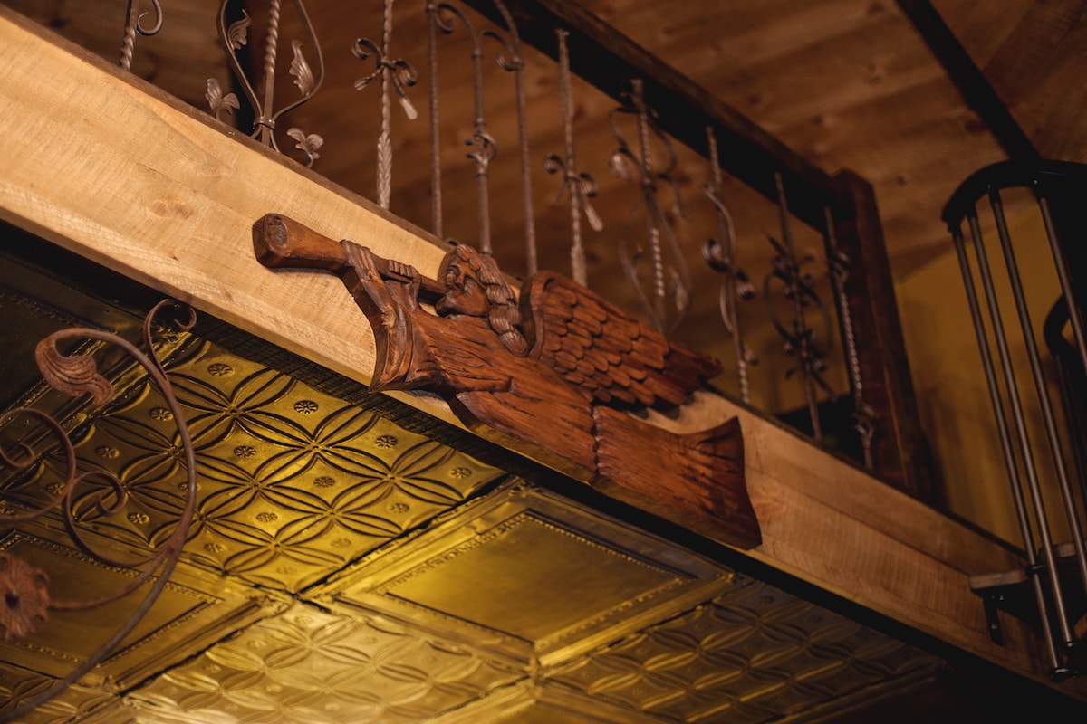 A detailed wooden carving of a cherub playing a trumpet is displayed on a rustic wooden beam. The beam is adorned with intricate ironwork, and an antique tin ceiling provides a rich texture beneath. The warm lighting enhances the craftsmanship of this unique feature.
