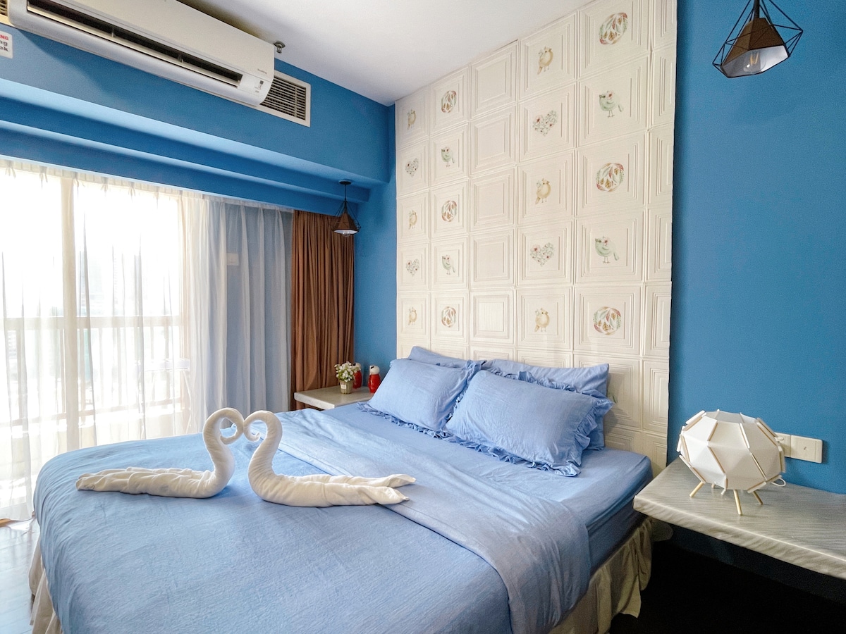 A spacious bedroom features blue walls and a decorative feature wall. A large bed is adorned with soft blue linens and two towels shaped like swans. Natural light filters through sheer curtains, illuminating a modern bedside table and pendant light.