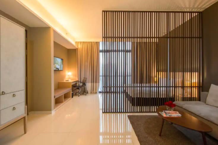 A modern room features a stylish partition creating a cozy separation between the sleeping area and lounge. A spacious bed is visible beyond the divider, while a small desk and chair are positioned under a wall-mounted television. Soft lighting enhances the contemporary decor.