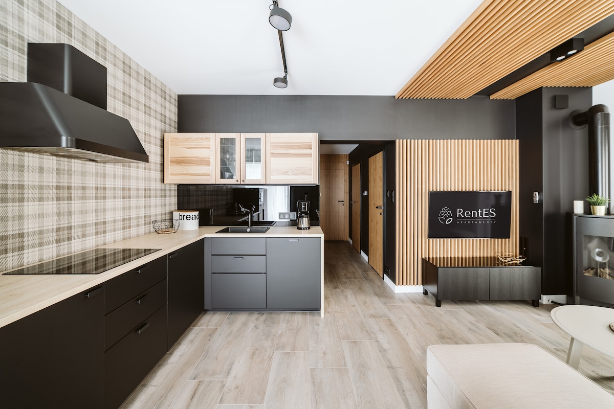 The kitchen area features modern cabinetry with a blend of light wood and matte black finishes. A sleek stovetop and ample counter space provide functionality, while decorative items add a touch of style. The adjacent living area is subtly visible, showcasing a cohesive design.