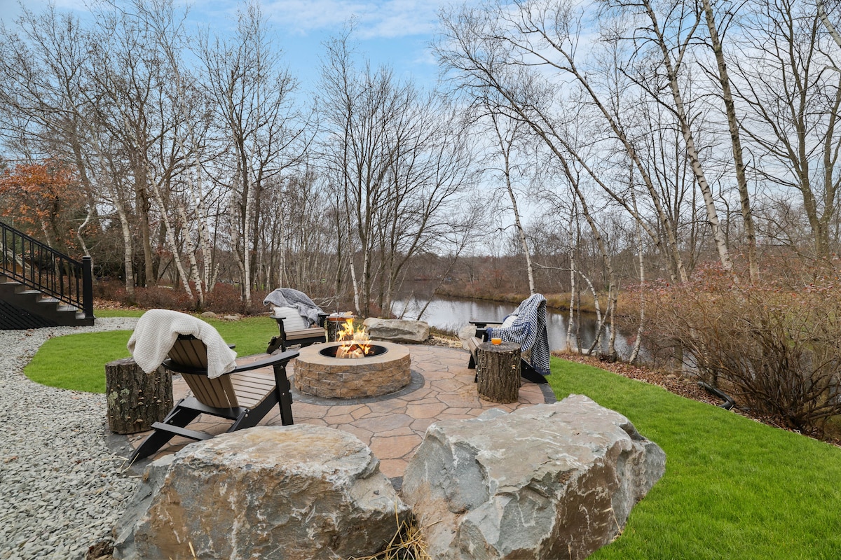 A fire pit area is artfully arranged with wooden seating around a stone circle. The surrounding landscape features a pristine lake and bare trees, creating a serene setting. Cozy blankets are draped over the chairs, inviting relaxation by the fire.