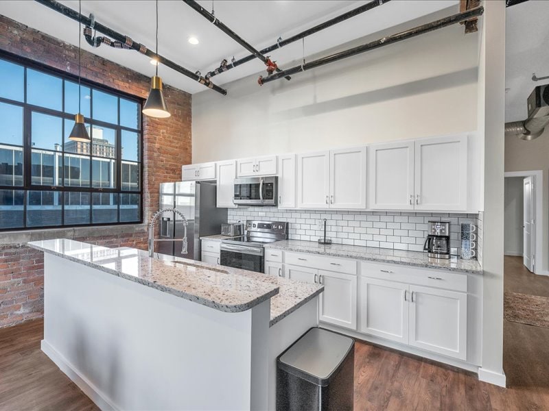 A modern kitchen features a large island with a speckled countertop and seating. Stainless steel appliances, including a microwave and stove, are complemented by white cabinetry. Natural light flows through expansive windows, which highlight the rustic brick wall.