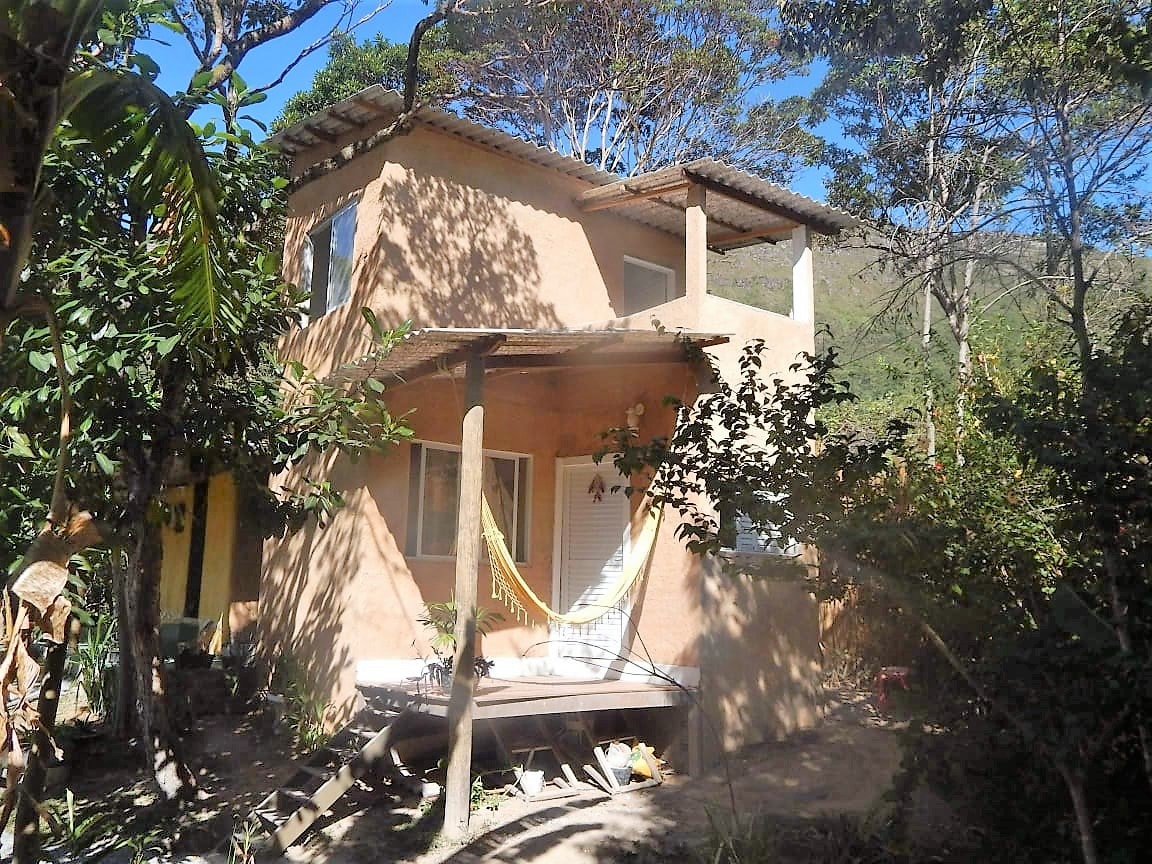 A two-story building is nestled among trees, exhibiting a natural earth-tone exterior. A small porch with a hammock invites relaxation. Windows allow natural light to brighten the interior, while the surrounding greenery enhances the tranquil setting.