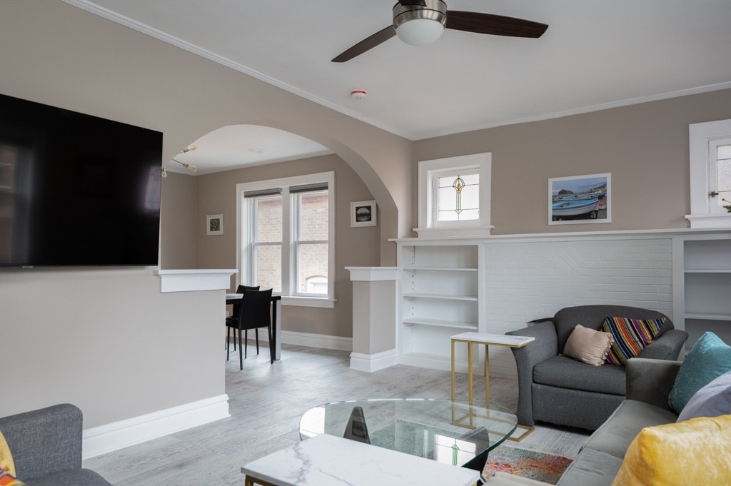 A modern living area features a comfortable gray sofa and armchair, complemented by colorful cushions. A glass coffee table sits centrally, reflecting a wall-mounted television above. Natural light floods through the three nearby windows, highlighting the light gray flooring and neutral wall tones.