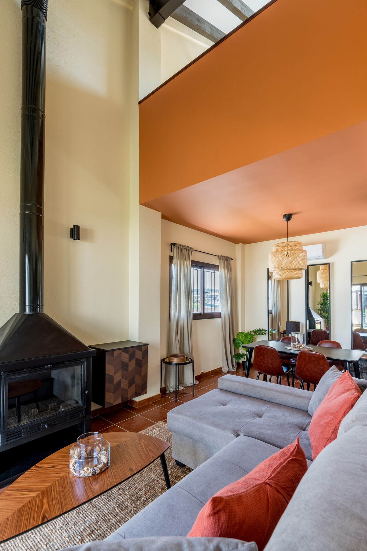 A cozy living area is highlighted by a modern gray sofa and a decorative coffee table. An ornate fireplace adds warmth to the space. Large windows provide natural light, while an orange ceiling and stylish light fixtures enhance the interior's contemporary design.