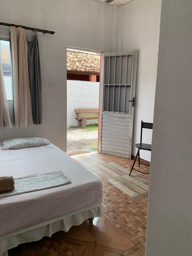 A simple yet cozy room features a spacious bed with a light coverlet. A chair is positioned nearby, and the partially open door leads to an outdoor area. Natural light floods in from the window, contributing to a bright and airy atmosphere.