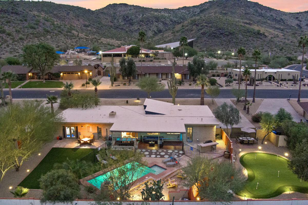 An aerial view showcases the home against a mountainous backdrop. The landscaped yard features a heated pool and a putting green, surrounded by desert vegetation. Outdoor seating areas are positioned near the pool, while the house displays unique architectural lines.