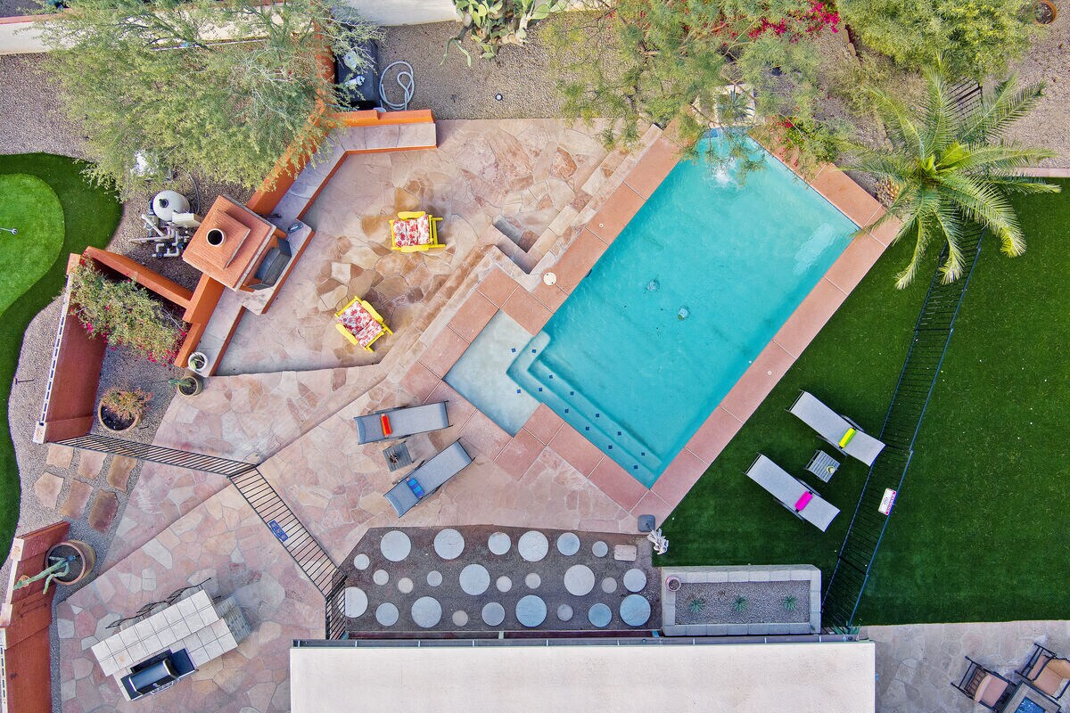 An aerial view showcases a landscaped backyard featuring a heated pool, lounge chairs, and a spacious patio area. Vibrant seating arrangements and a putting green are visible, along with lush greenery framing the area, creating an inviting space for relaxation and entertainment.