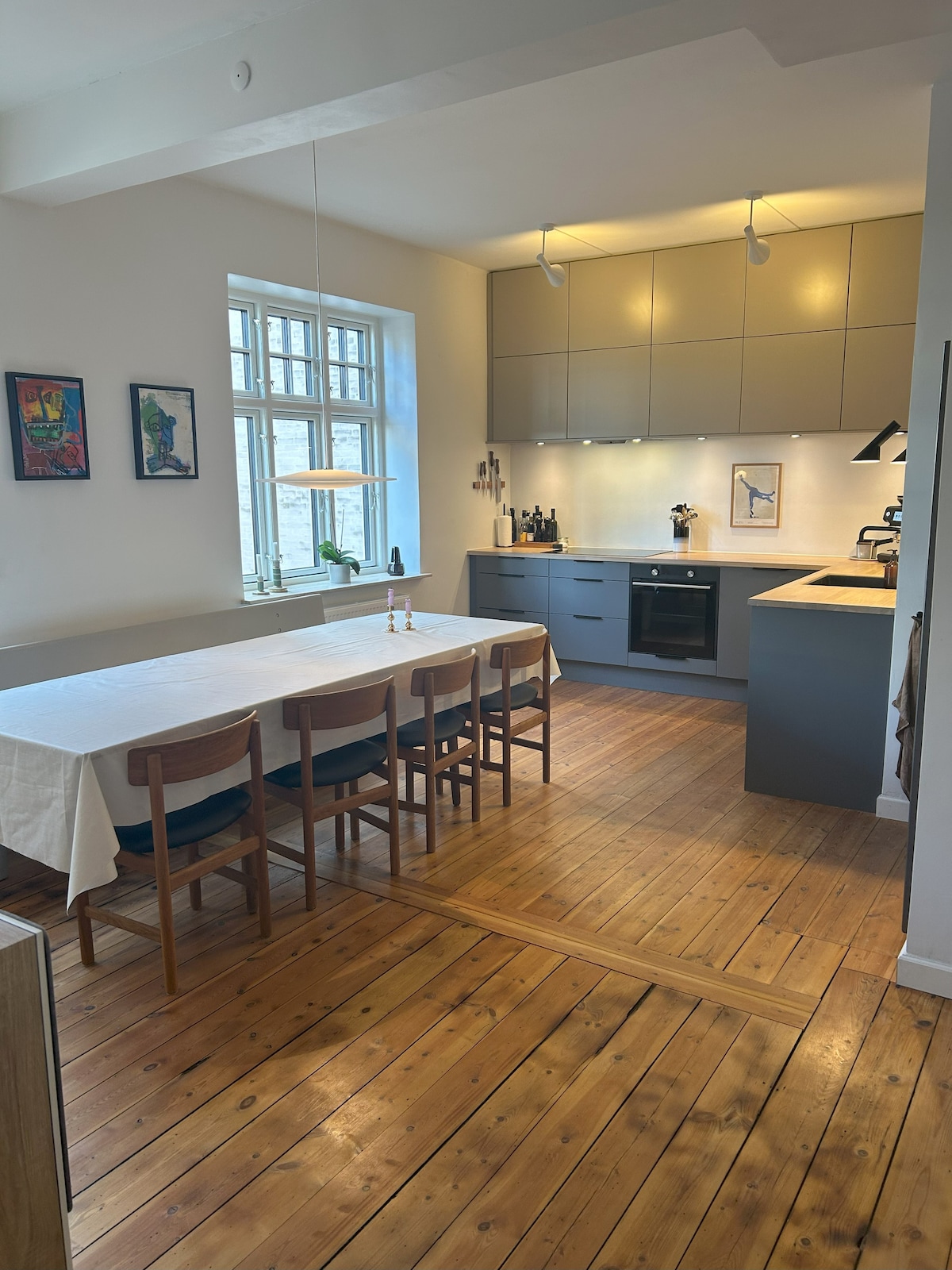A spacious kitchen and dining area are featured, with a long dining table and several wooden chairs arranged. Modern cabinetry and a sleek oven highlight the kitchen's functionality. Natural light floods through a large window, complementing the warm wood floors.