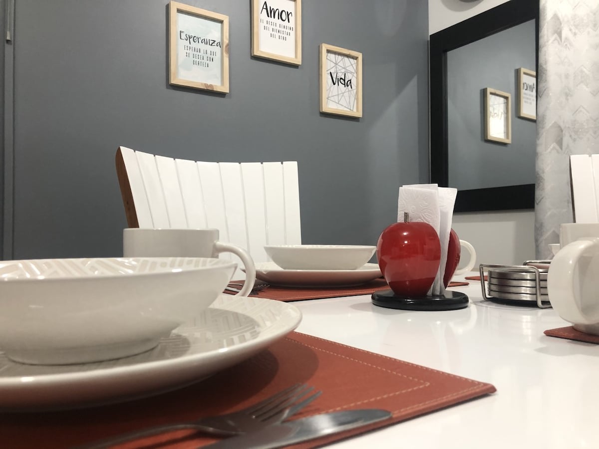 A dining table is set with white dishware, including bowls and cups, placed on a textured placemat. A red decorative apple and napkins are arranged centrally. Light-colored wooden chairs contrast with the gray wall featuring framed inspirational art. A mirror reflects the cozy dining area.