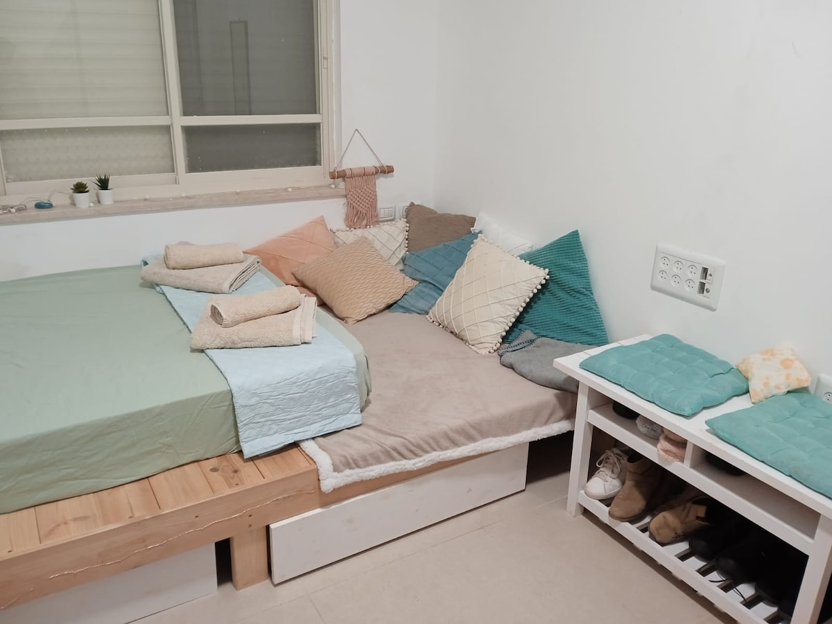 A sleeping area is arranged with a spacious bed layered in a light green coverlet. Cushions in various colors are placed along one side, providing comfort. A small white shelf holds shoes, while natural light enters through a large window adorned with minimal decor.