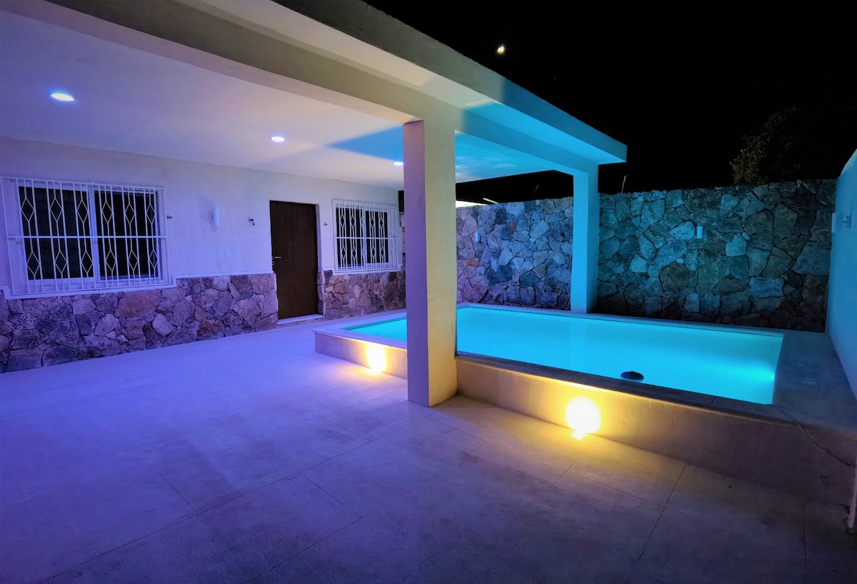 An outdoor area is illuminated by soft lighting, featuring a private pool and a spacious deck. A stone wall provides privacy, while large windows allow natural light into the indoor space. The serene atmosphere creates a perfect setting for relaxation.