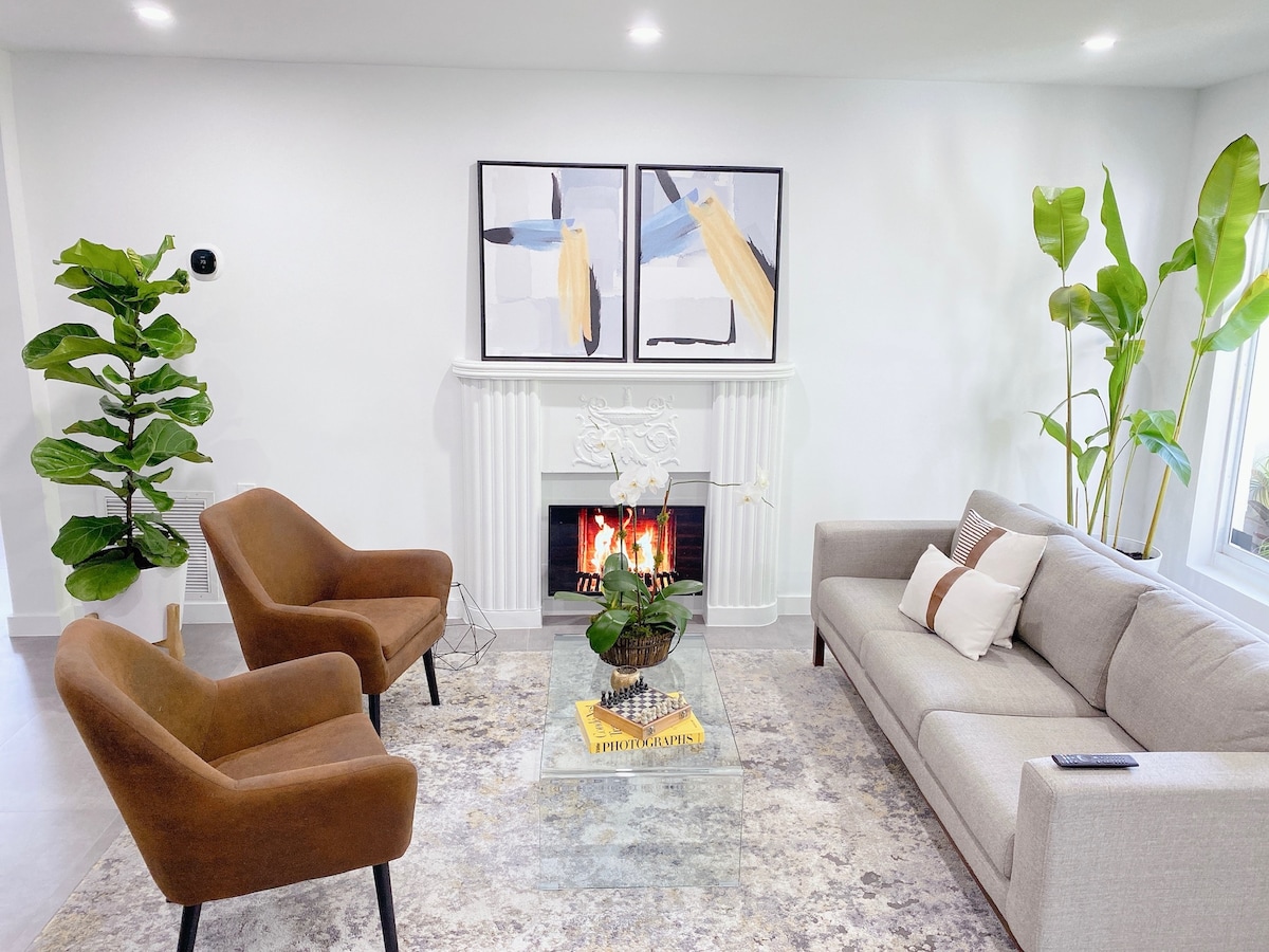 A well-lit living area features a cozy sofa and two armchairs, complemented by a decorative area rug. A fireplace serves as a focal point, with art pieces displaying a modern design above it. Lush indoor plants add greenery to the space.