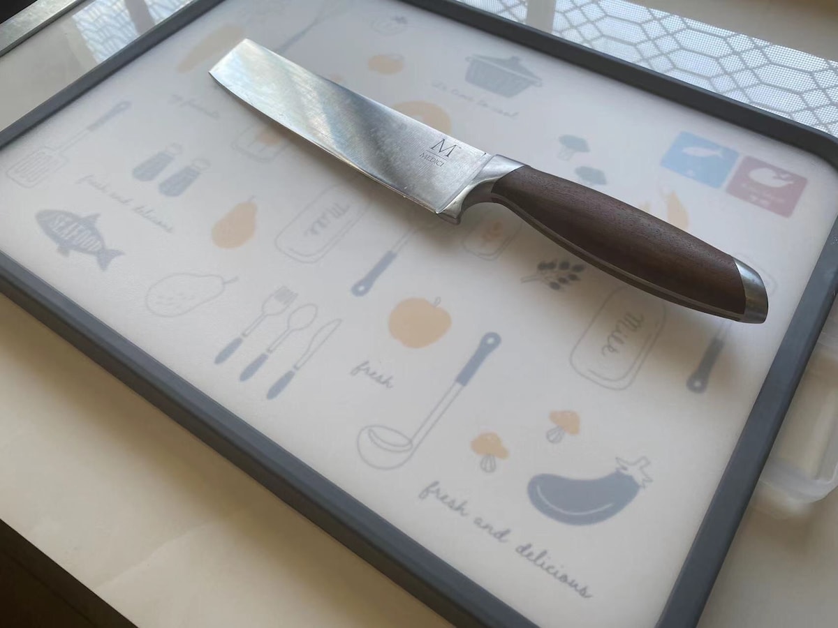 A kitchen countertop is depicted featuring a cutting board adorned with illustrated food items and utensils. A sharp kitchen knife with a wooden handle is positioned on the board, emphasizing its readiness for meal preparation.