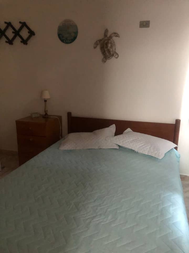 Apartmán 1 - spálňa s manželskou posteľou: posteľ, nočný stolík s lampou a malou škatuľkou, skriňa a malá polička na knihy, nástenné ozdoby, zatemnený záves, tienené okno.