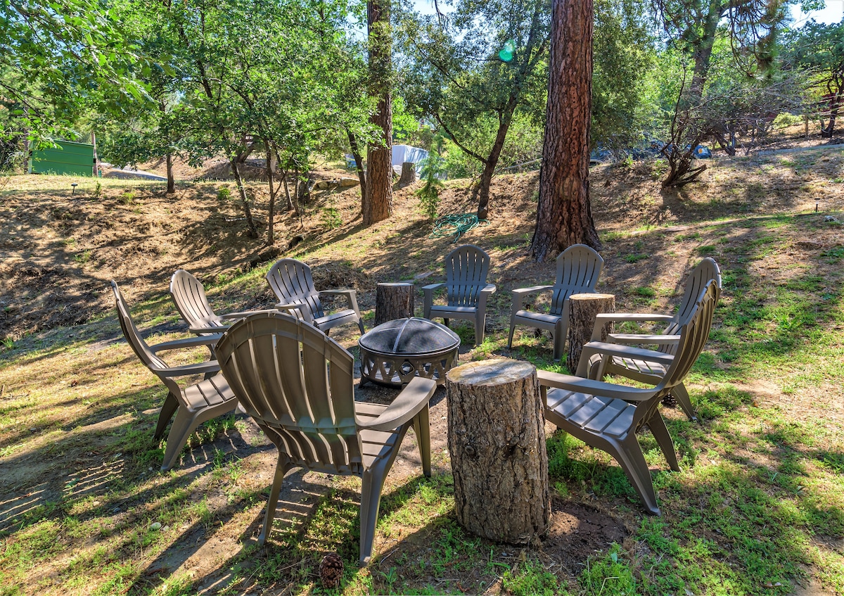 A circle of sturdy outdoor chairs is arranged around a central fire pit, situated on a grassy area surrounded by trees. The setting offers a peaceful atmosphere, with sunlight filtering through the foliage, creating a cozy spot for gathering.