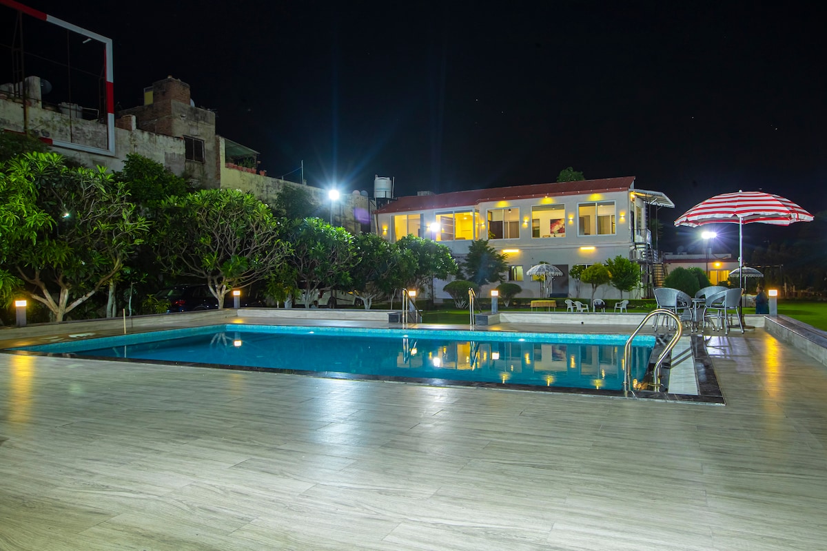 A serene pool area is illuminated by garden lights, reflecting a two-story building in the background. The pool is surrounded by well-maintained greenery, with several chairs arranged nearby. A striped umbrella offers shade, enhancing the tranquil evening ambiance.