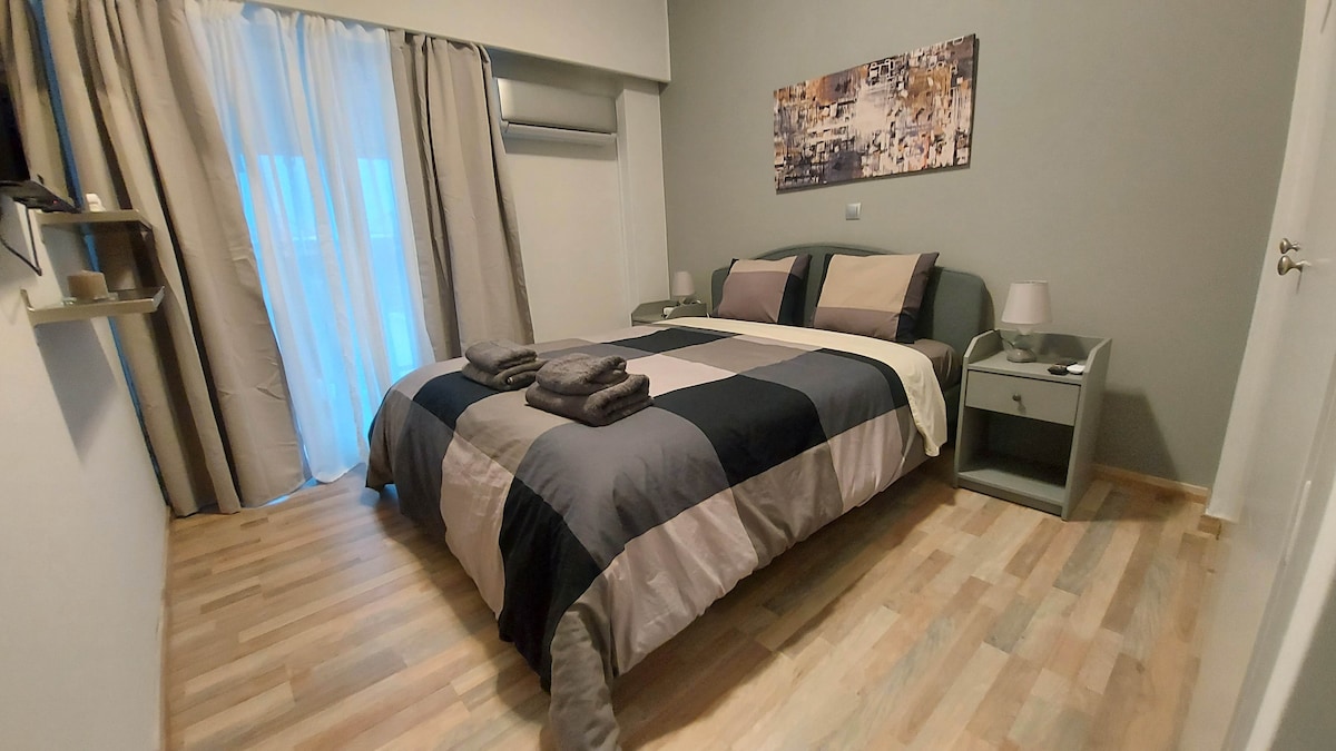 A spacious bedroom features a large bed with a patchwork-style duvet and neatly arranged towels on top. Nightstands on either side of the bed are complemented by lamps. Light filters through sheer curtains, and wood flooring adds warmth to the room.