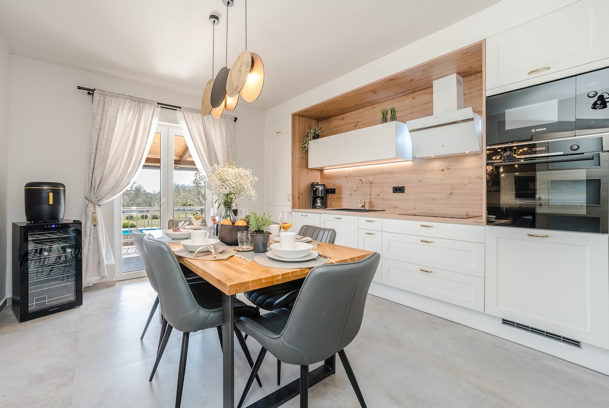 A modern dining area is featured, showcasing a wooden table set for four with stylish black chairs. The light wooden cabinetry complements the space, and large glass doors invite natural light, providing views of the outdoor area. A fridge and coffee maker are visible in the kitchen.